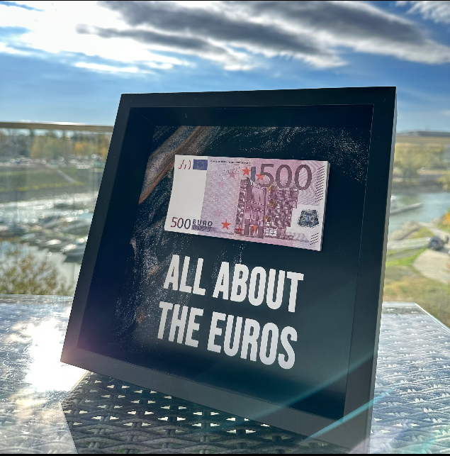 "ALL ABOUT THE EUROS" feliratú 3D képkeret 500 eurós bankjeggyel, kültéri üvegasztalon, napsütésben.