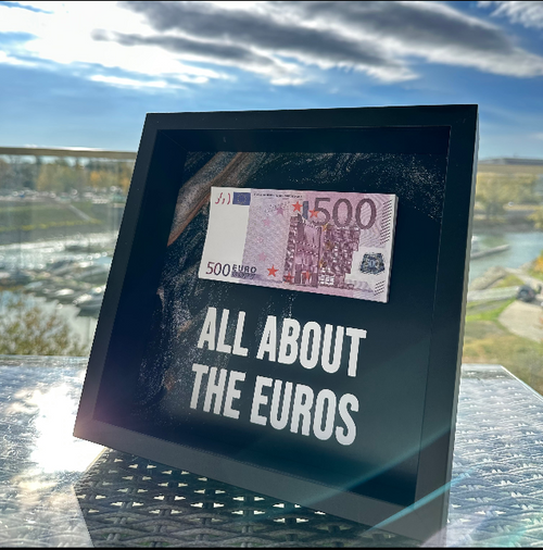 "ALL ABOUT THE EUROS" feliratú 3D képkeret 500 eurós bankjeggyel, kültéri üvegasztalon, napsütésben.