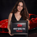 Egy mosolygós barna hajú nő egy fekete képkeretet tart, amelyben egy piros 3D-s autókulcs és fehér, illetve piros betűkkel a "NEED MONEY FOR FERRARI" felirat látható. A háttérben egy elmosódott piros sportautó parkol.